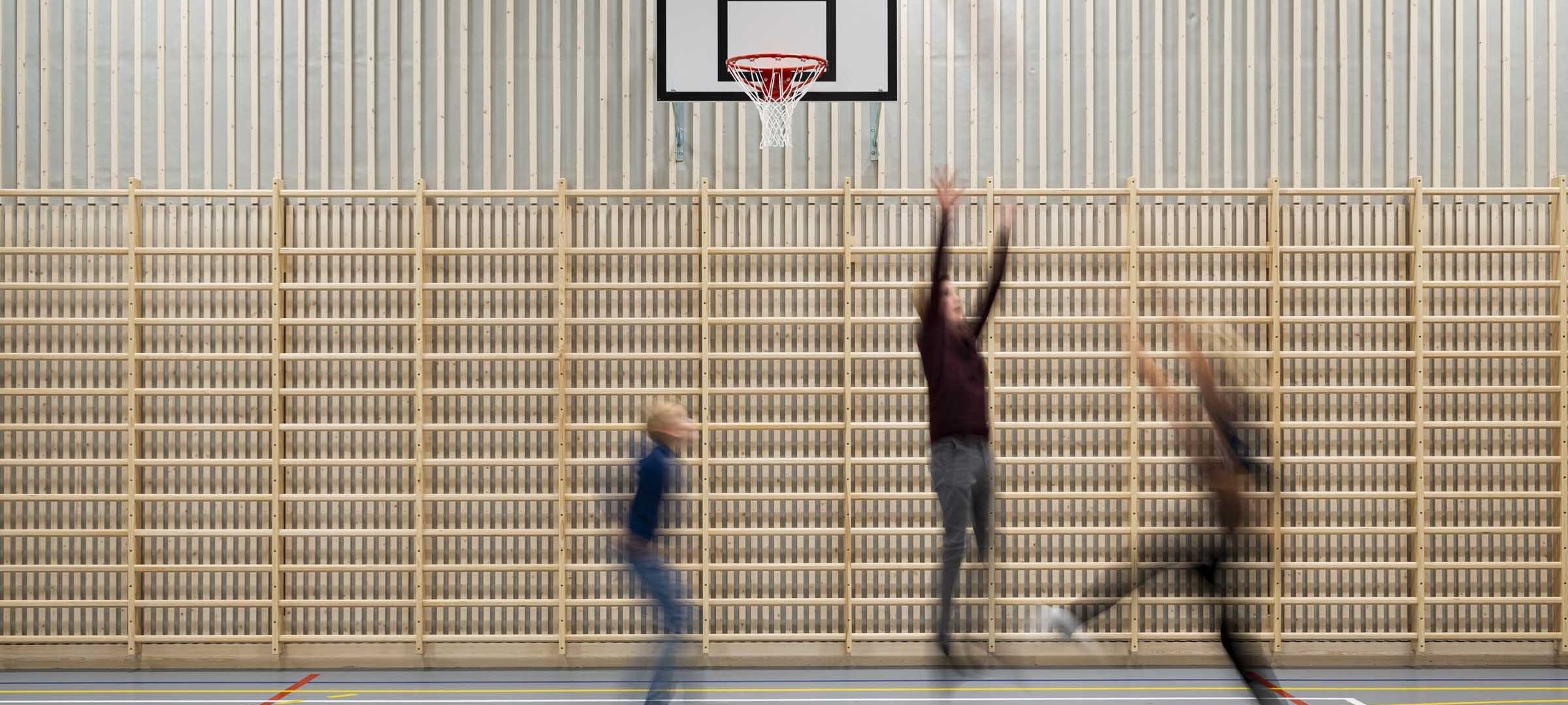 Tre personer spiller basketball i gymsal i forkant av en ribbevegg som er plassert langs hele den ene veggen. De hopper opp for å kaste ballen i kurven. Foto.